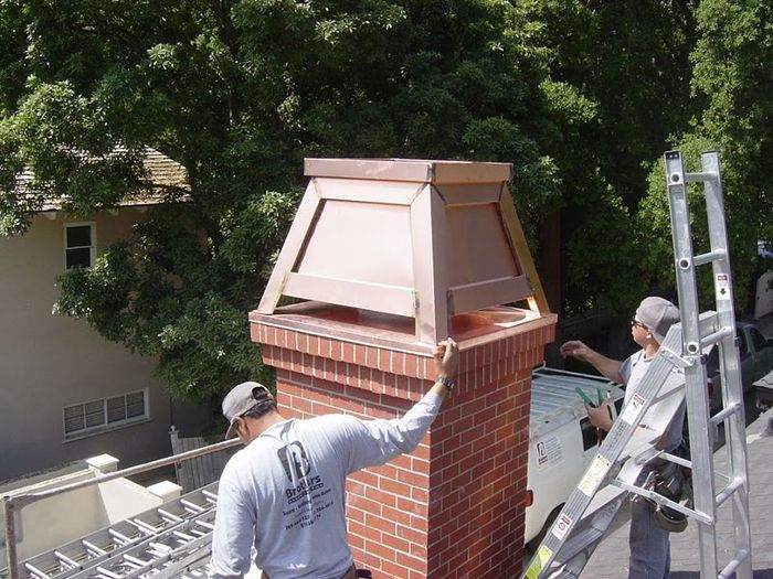 Two workers install a copper chimney cap on a brick chimney on a rooftop.