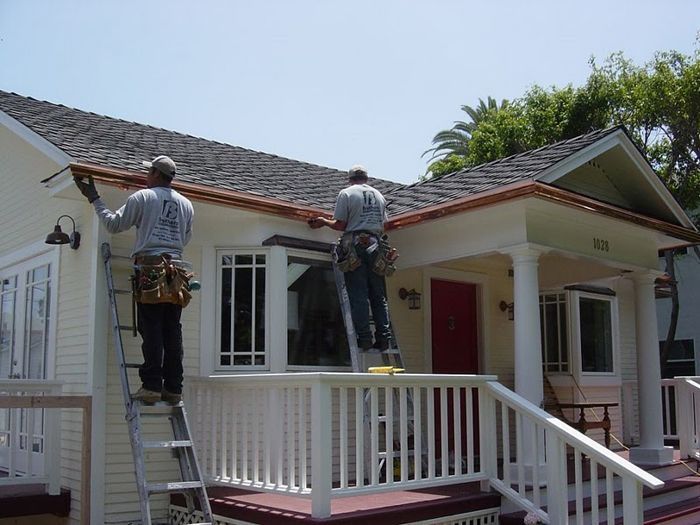 Two workers installing copper gutters on a light-colored house with a red door.