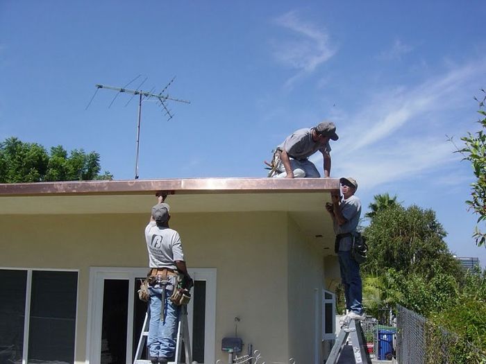 Three workers installing copper gutters on a house roof under a blue sky.