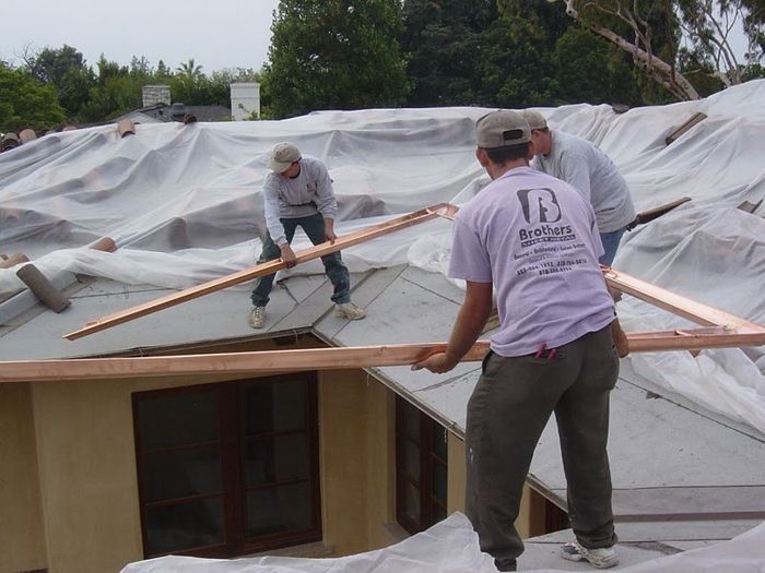 Three workers on a roof installing copper flashing. House is covered with protective plastic.