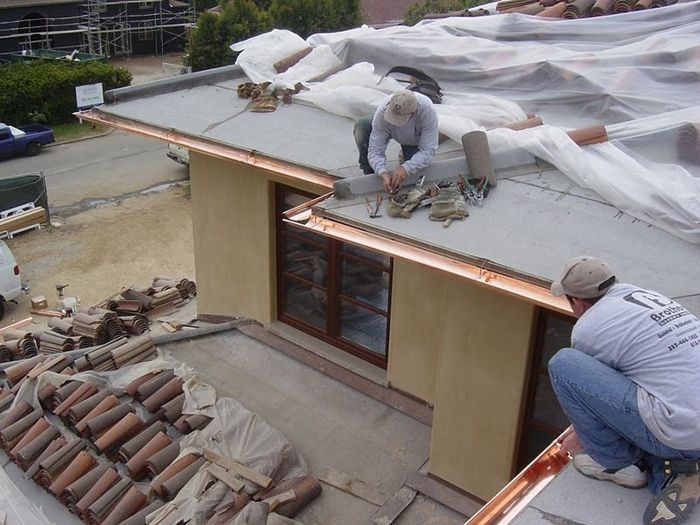 Two workers installing copper guttering on a building's roof. Brown tiles and tools scattered around.