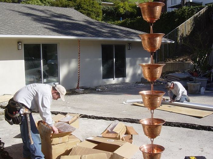 Men install copper rain chain near a house. Boxes of materials are on the ground.
