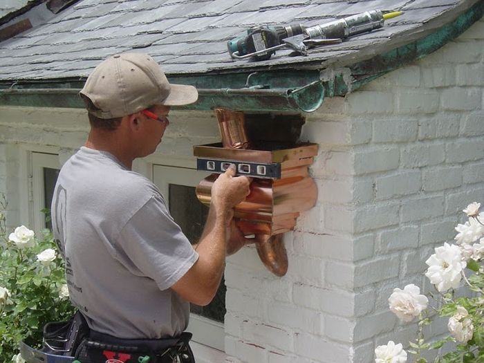 Man installing a copper downspout on a white brick building, green roof, sunny outdoor setting.