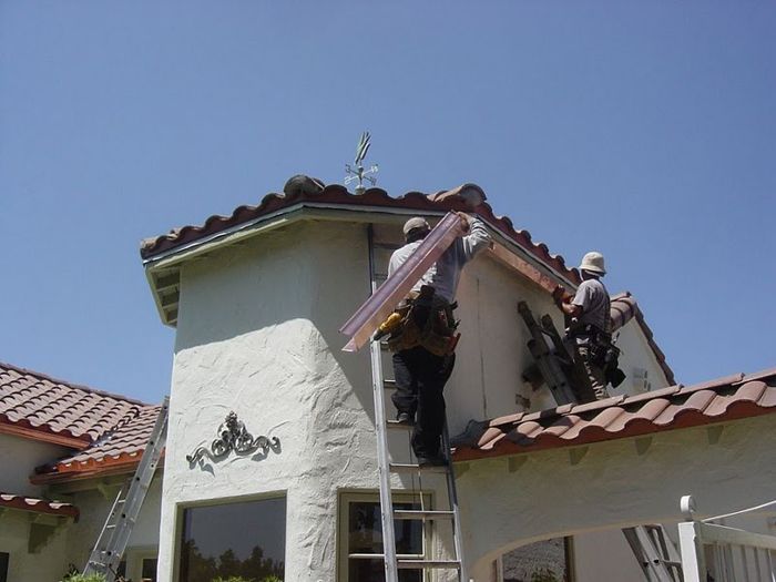 Two workers install gutters on a white stucco building with red tile roof, on a sunny day.