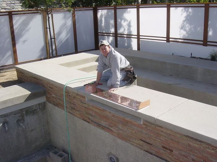Man working on a concrete pool deck. He's kneeling and placing a brick, setting is outdoors, near a white fence.