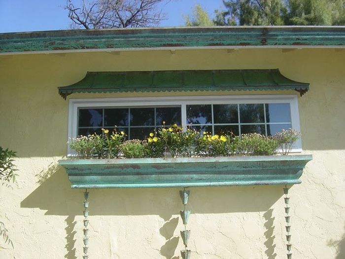 Green-roofed window with a planter box filled with yellow flowers on a yellow stucco wall.