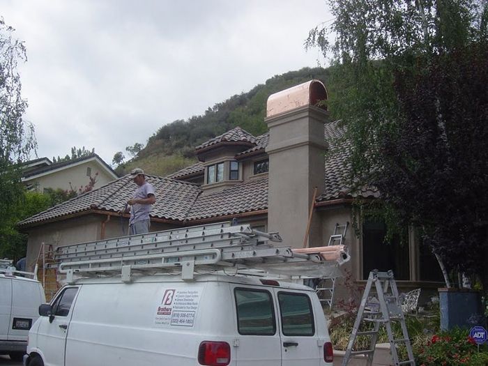 Man on a chimney, working with copper. White van in front. House with a tiled roof, mountain in the background.