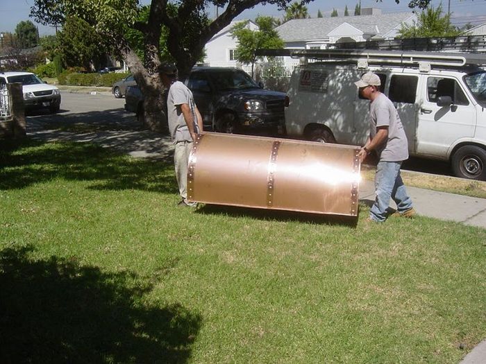 Two men carrying a large, copper-colored cylindrical object across a green lawn. A white van is parked nearby.