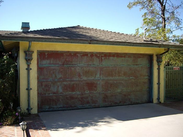 Rusty garage door in front of a yellow stucco building with a brown roof.