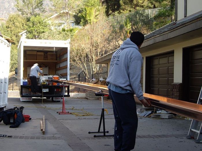 Two workers carrying copper gutter near truck and house.