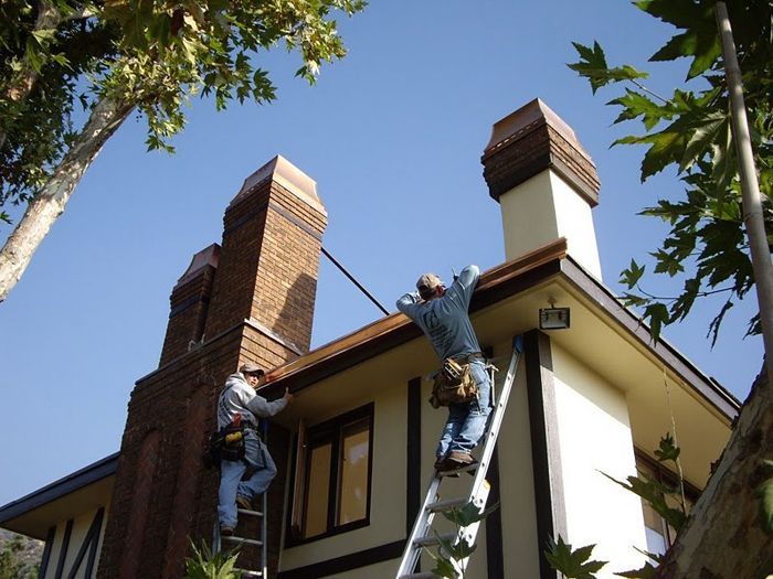 Two men on ladders working on a house roof near brick chimneys on a sunny day.