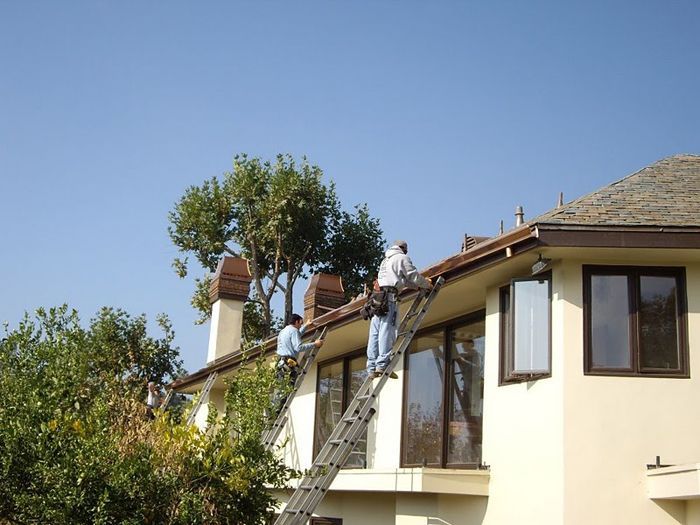 Workers on a roof, using ladders, repairing roof tiles on a two-story beige house with brown trim on a sunny day.