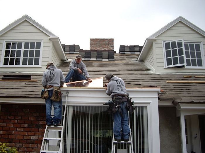 Three roofers on a house roof removing old shingles. Brick chimney, windows visible. Overcast day.