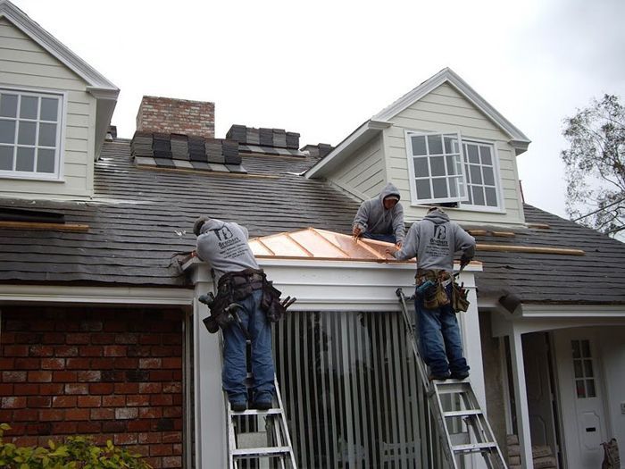 Three workers on ladders install copper roofing trim on a house with dormer windows and brick details.