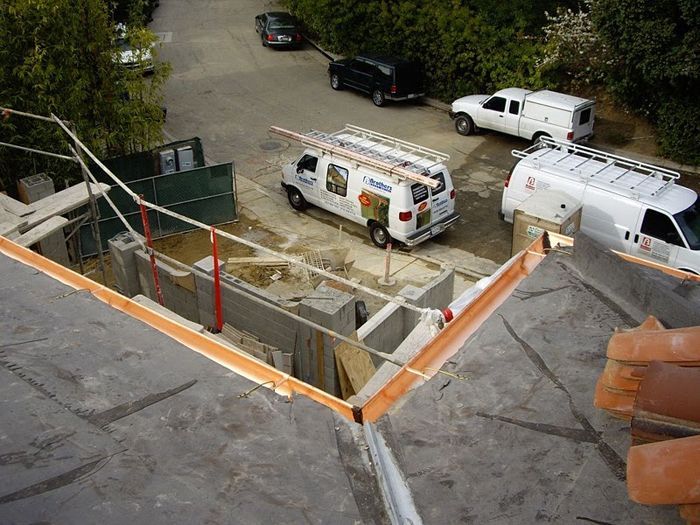 Construction site: workers on a roof, vehicles parked on the street. Safety barriers around the edge.
