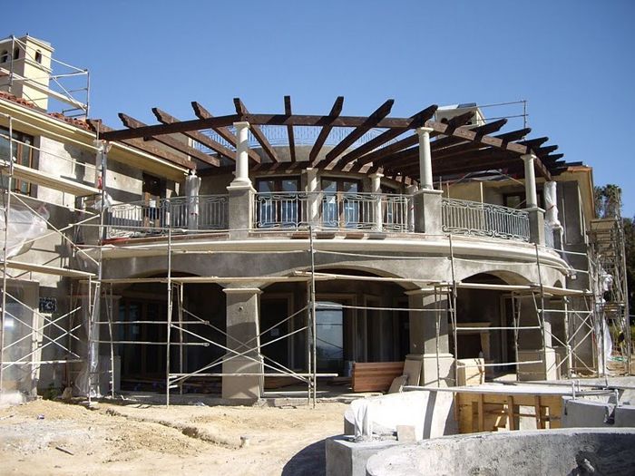 Large, ornate house under construction; scaffolding surrounds a balcony with a pergola.
