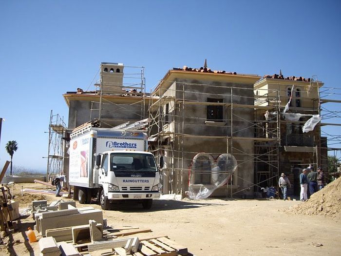 Construction site: truck parked, partially built two-story house, scaffolding, workers, blue sky.