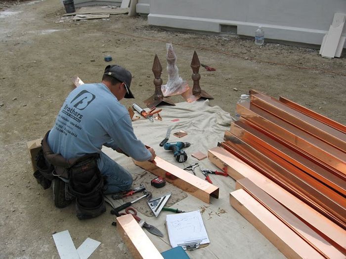 A construction worker kneels on a tarp, assembling copper beams with tools and finials in an outdoor setting.