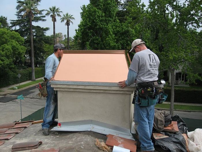 Two roofers installing copper flashing on a chimney on a tile roof.