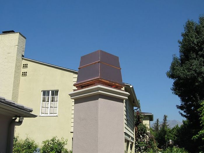 Copper chimney cap on a beige brick chimney against a blue sky and a yellow building.