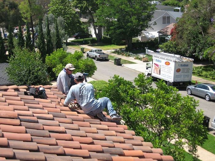 Two roofers on a clay tile roof; street with truck and cars in the background.