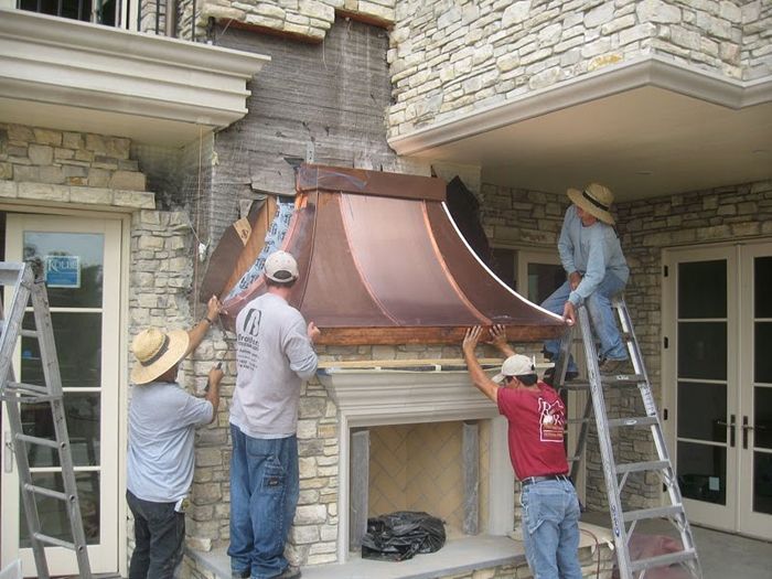 Four workers installing a copper fireplace hood on a stone building exterior.