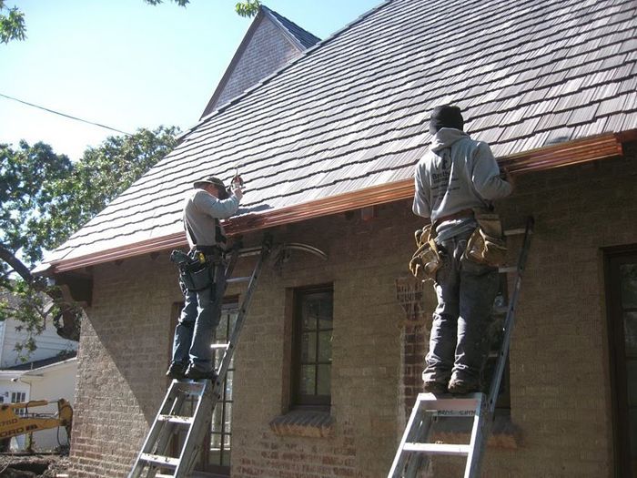 Two workers on ladders installing copper gutters on a brick house with a shingle roof.