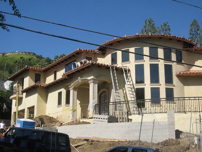 Two-story house under construction, beige stucco, arched windows, red tile roof, ladder, blue sky, construction vehicles.