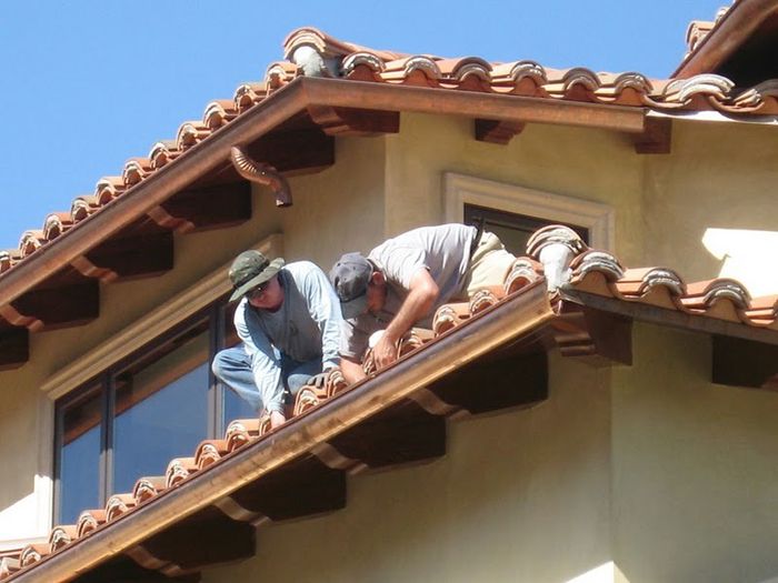 Two workers on a roof, installing brown tiles on a building with tan stucco and blue sky.