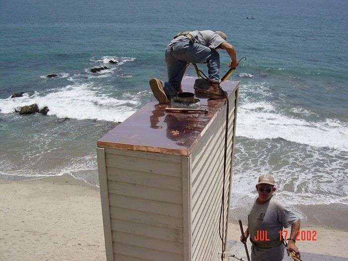 Two men working on a chimney with ocean waves in the background. One man on top, the other below.