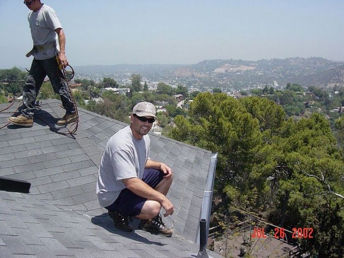 Two roofers on a rooftop overlooking a valley. One squats, smiles; the other works. Sunny day.