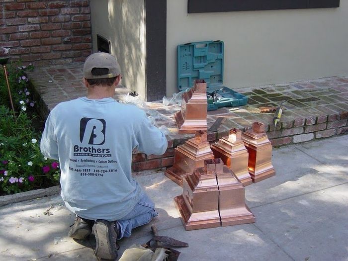 Man kneeling, working with copper decorative elements near a brick structure. Sunny outdoor setting.
