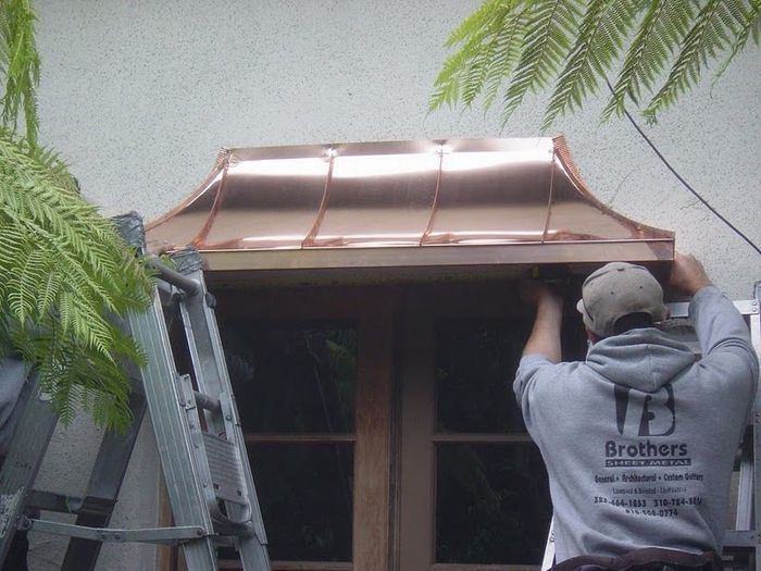 Man installing a copper awning above a window. The setting is outdoors with a ladder.