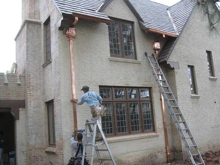 Two men installing copper gutters on a two-story stucco house with a ladder.