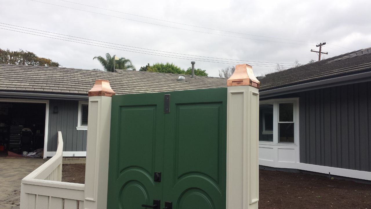Green gate with copper-topped pillars in front of a gray house, cloudy sky.