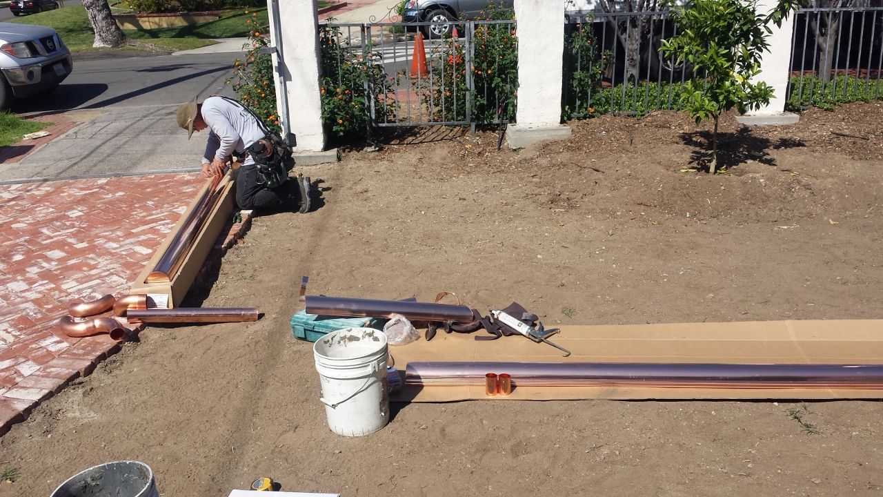 Man installs copper pipe, tools and supplies on ground. Outside near sidewalk and gate.