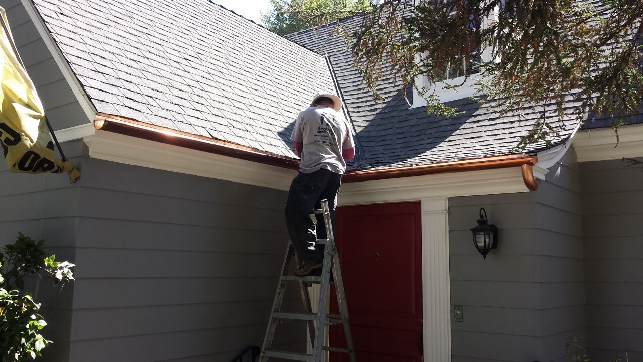 Person on a ladder installing copper gutters on a gray house with a red door.