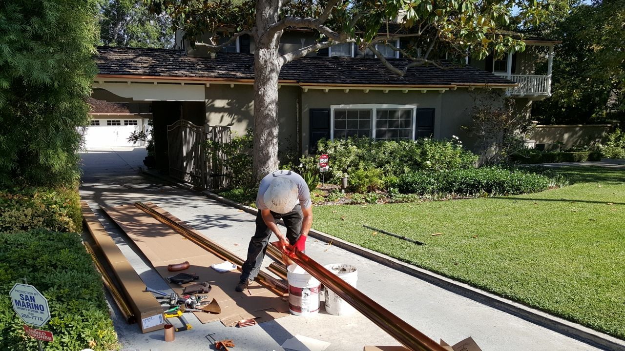 A man in a white hat cuts lumber on a driveway in front of a house with grass and a tree.