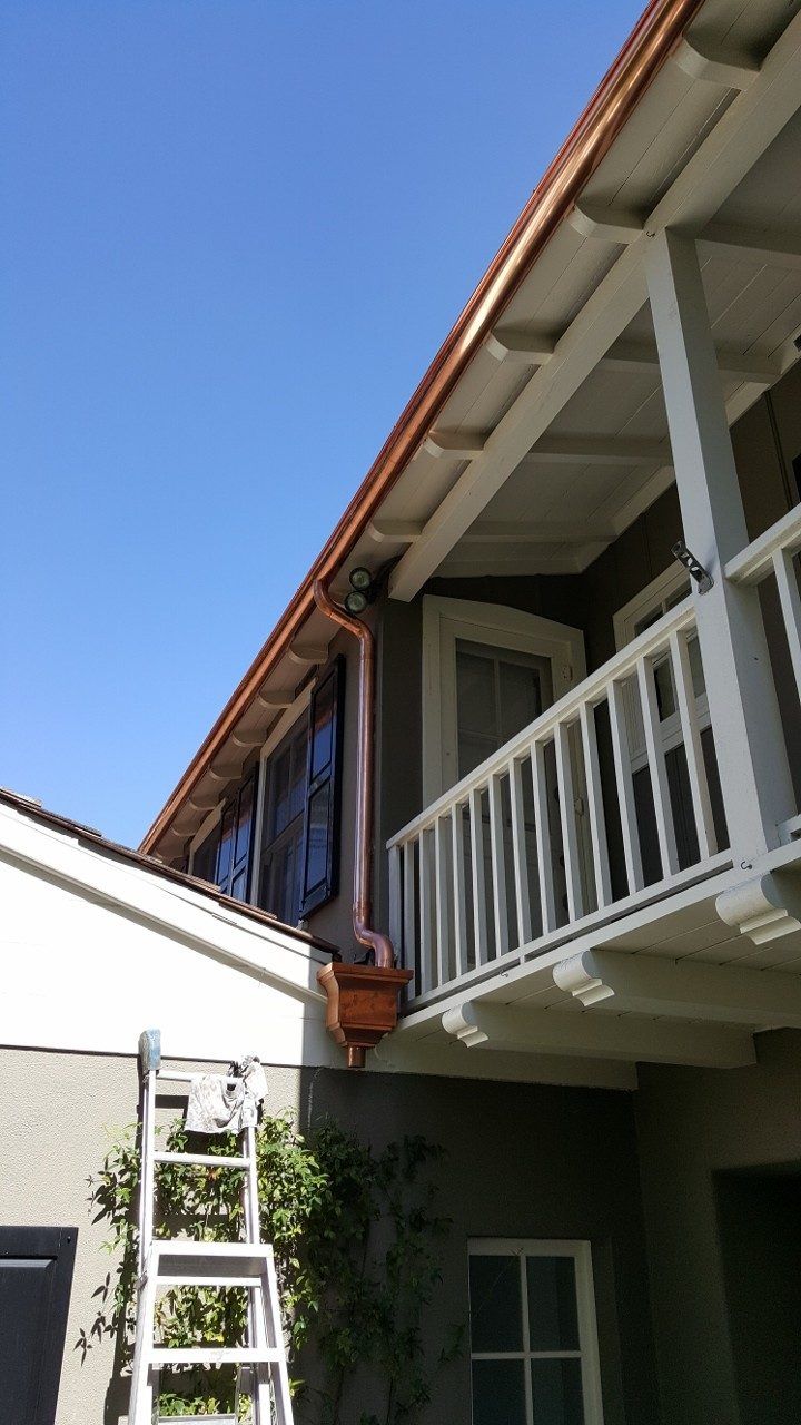 Copper gutters on a two-story house with a balcony, under a blue sky; ladder leans against the wall.