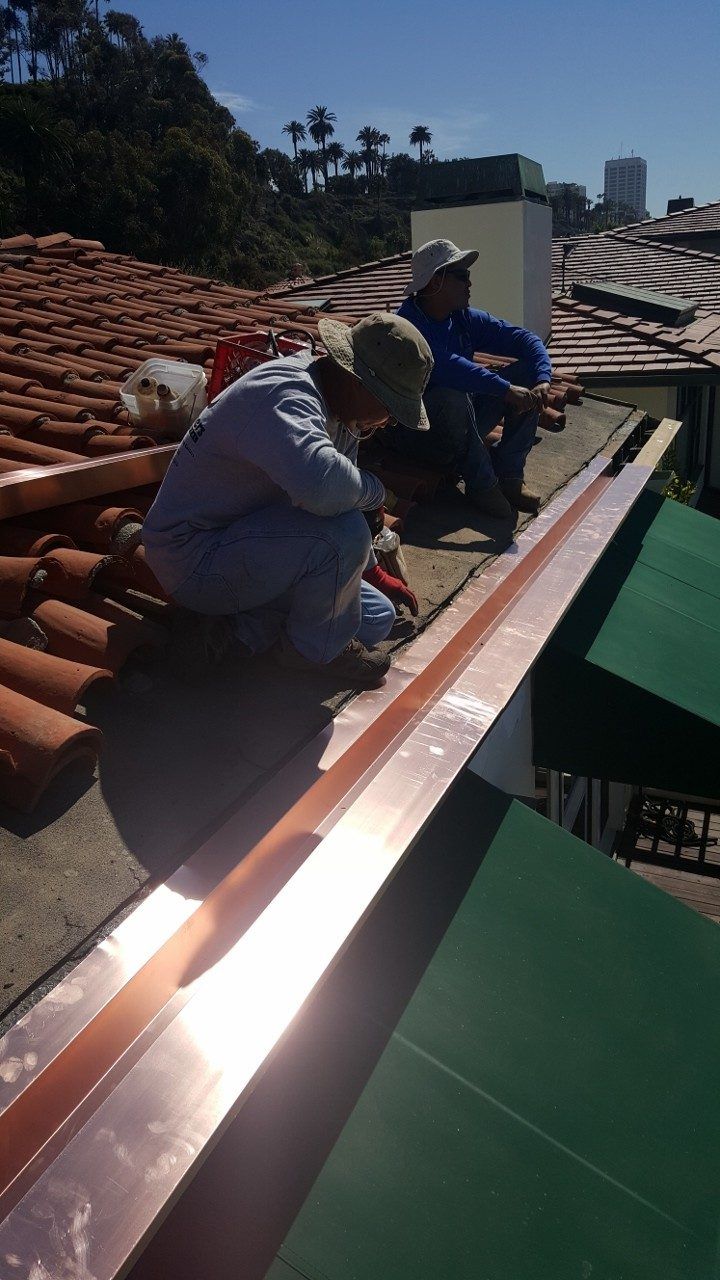 Two roofers install copper flashing on a terracotta tile roof, sunny day.