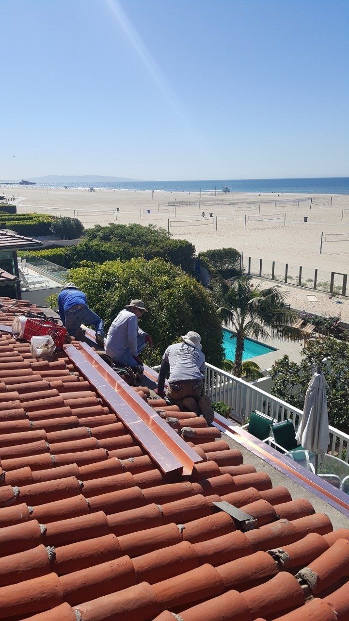 Workers installing copper flashing on a red-tiled roof overlooking a sunny beach.