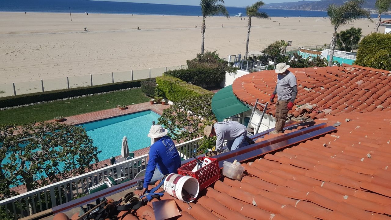 Three workers repairing a red tile roof overlooking a beach and a pool.