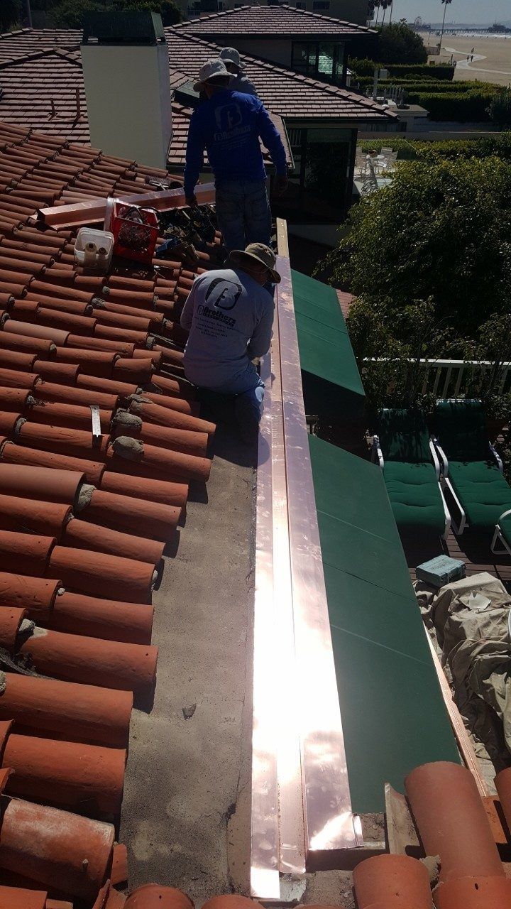 Two roofers installing copper flashing on a terracotta tile roof near a building with green awnings.