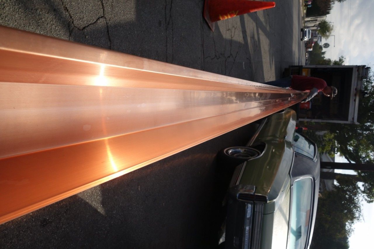 Long copper gutter being unloaded from a truck on a street. A person in a red shirt is handling the material.