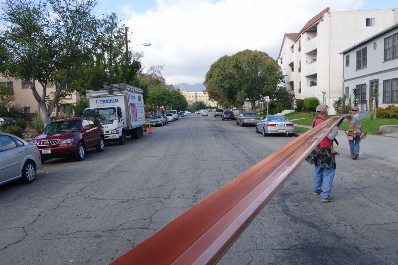 Two workers carrying a long, brown metal beam down a residential street.