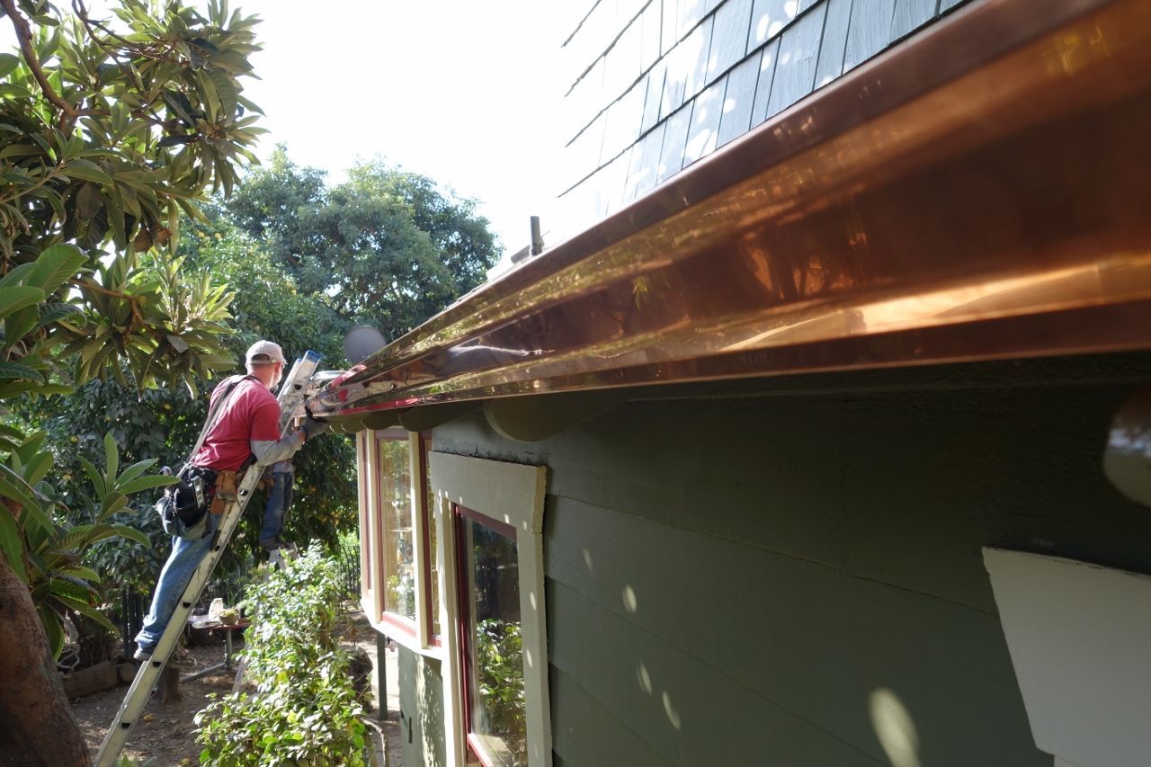 Man on a ladder cleaning a copper gutter on a green house.