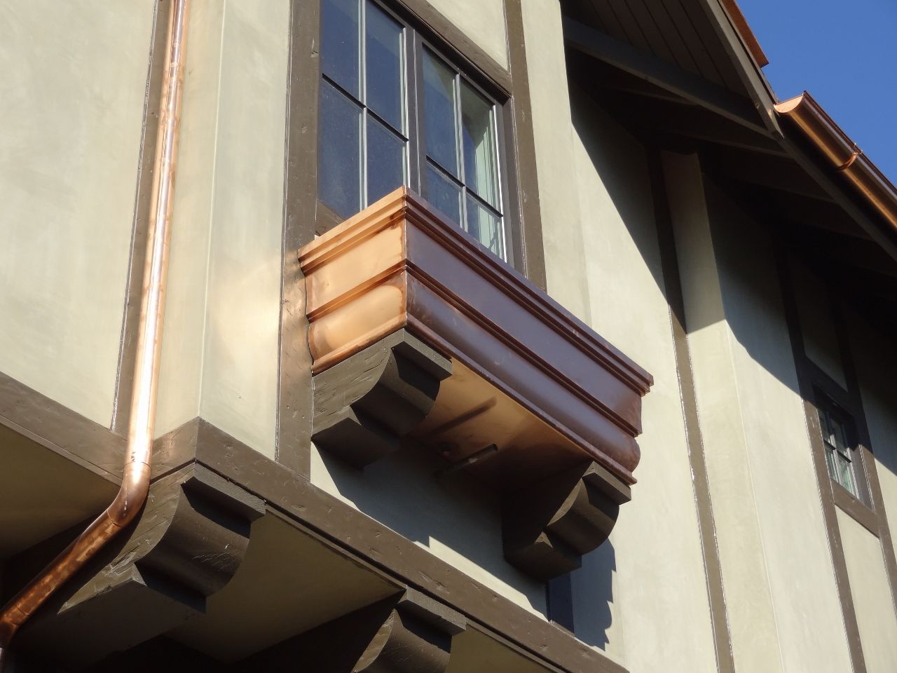 Copper window box on a tan and brown building. A copper gutter runs down the side.