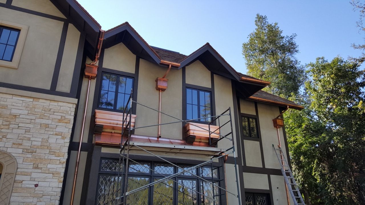 Two-story house exterior with copper gutters, dark brown trim, tan stucco, and blue windows. Scaffolding in front.