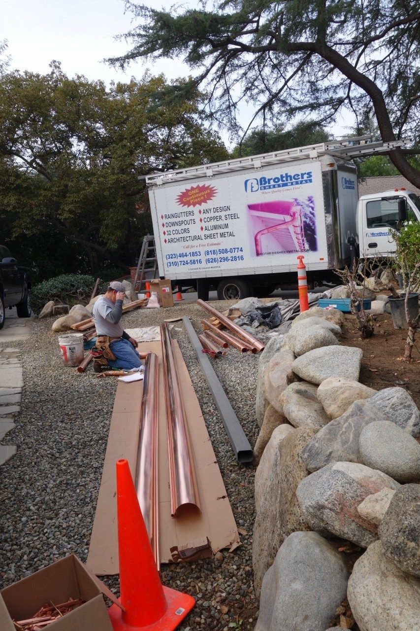 A worker installs copper tubes near a stone wall and truck. Tools and supplies are present outdoors.