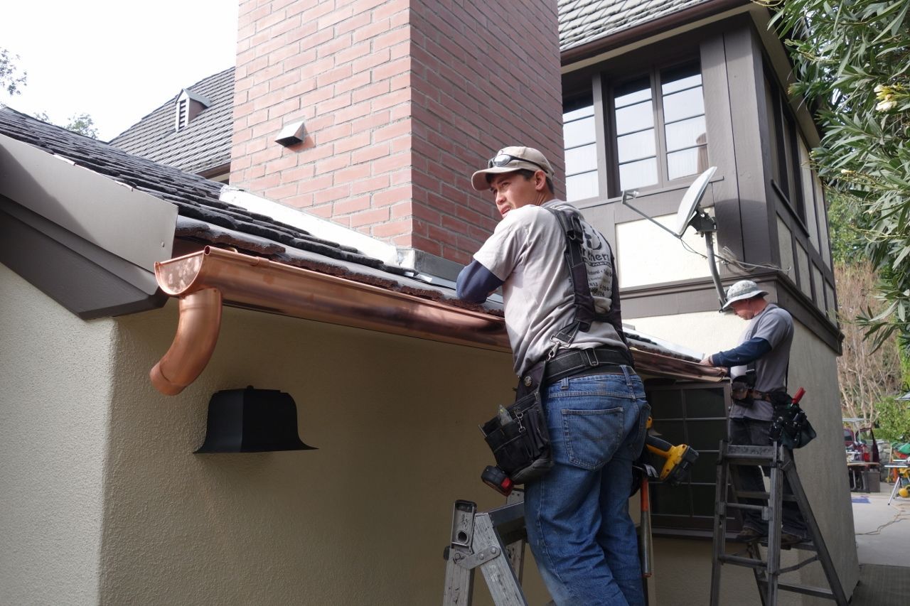Two workers installing copper gutters on a house near a brick chimney.